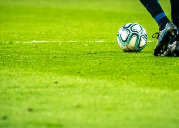 Çim sahada futbol topu ve krampon giymiş futbolcu ayakları.
