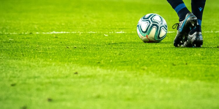 Çim sahada futbol topu ve krampon giymiş futbolcu ayakları.