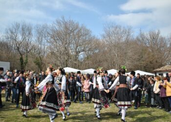 Geleneksel kıyafetler giymiş halk dansı ekibi açık havada el ele tutuşarak dans ediyor.