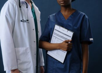 Laboratuvar önlüğü giyen doktor ve mavi üniformalı sağlık personeli duruyor.