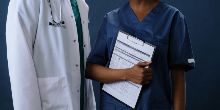 Laboratuvar önlüğü giyen doktor ve mavi üniformalı sağlık personeli duruyor.