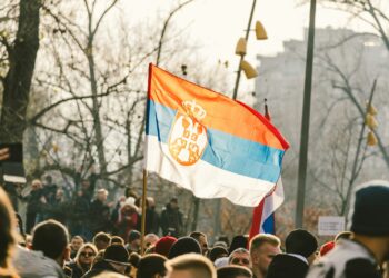 Sırbistan bayrağını sallayan protestocu kalabalık, arkada binalar ve ağaçlar.
