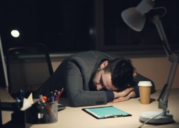 Loş ofis ortamında masaya kapanarak uyuklayan, yorgun görünümlü bir çalışan.