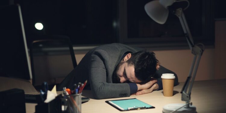 Loş ofis ortamında masaya kapanarak uyuklayan, yorgun görünümlü bir çalışan.